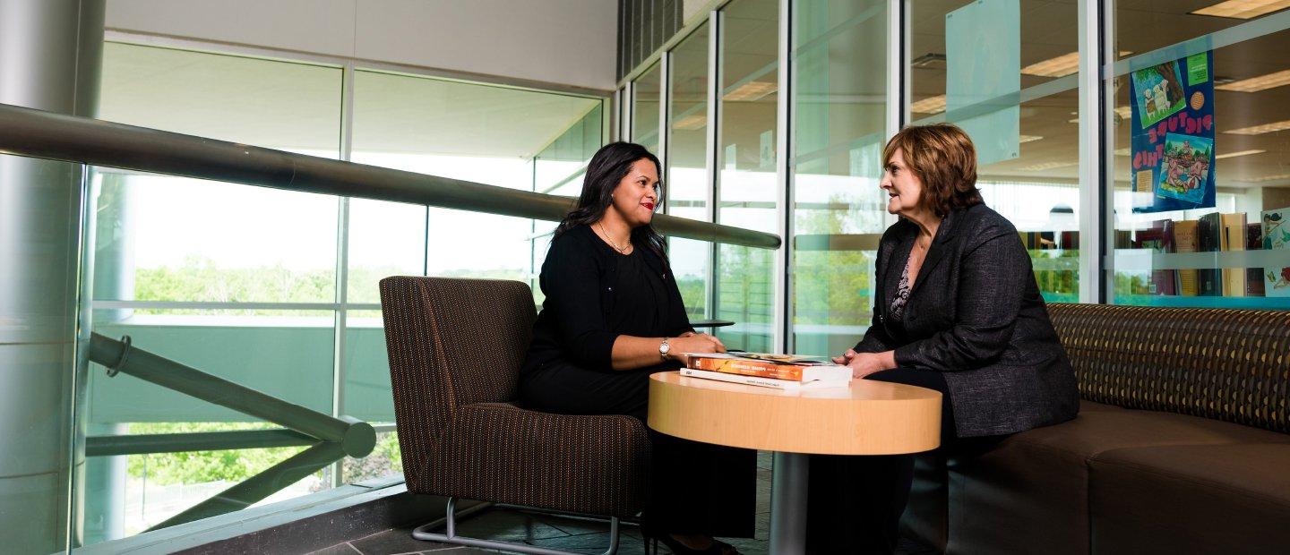 two women seated at a round table with books on it, windows behind them